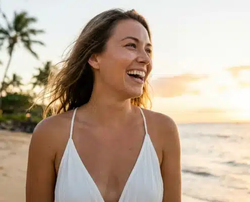 A smiling woman with glowing, youthful skin on a sunny Hawaii beach, representing the natural results of facial rejuvenation at Hawaii Facial Plastic Surgery.